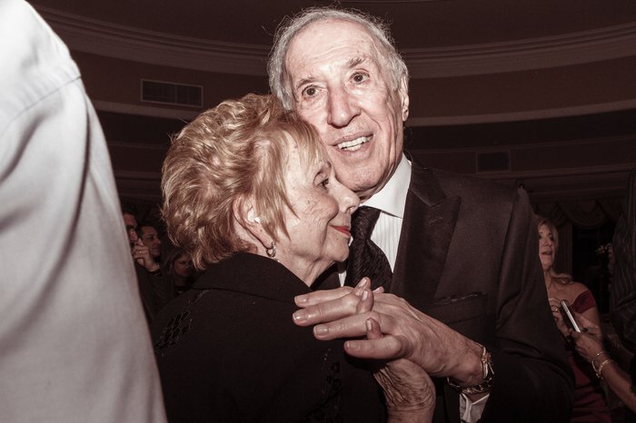 An elderly couple smile whilst dancing closely together in a crowd.