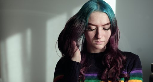A young woman with her eyes cast down, looking thoughtful. Her long hair is multi-coloured and she wears a rainbow sweater.