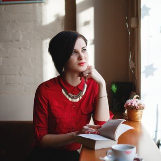 A young woman sits in a cafe with a book and a coffee, looking out of a window.