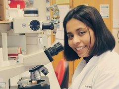 Anndita Roy - a researcher - in a lab looking directly at the camera with a microscope