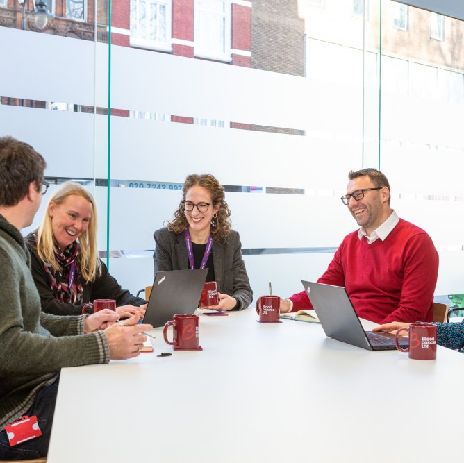 A group of people from the Blood Cancer UK executive board sitting around a table, laughing whilst having a meeting.