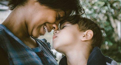 A smiling mother hugs her young son, who looks unhappy.