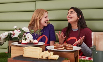 Two women wearing Blood Cancer UK t-shirts laugh together, sitting in front of some cakes.