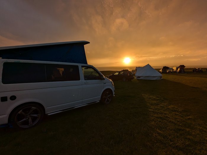 A campsite at sunset. In the foreground there is a white campervan with popped up top. Further away, a few tents and vehicles are also in the field.