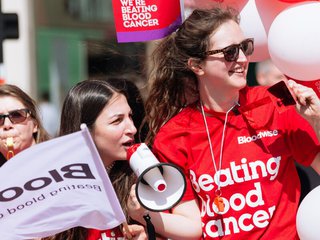 A group of Blood Cancer UK supporters stand with balloons and flags, cheering with a megaphone and whistles.