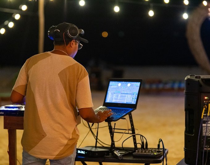 A DJ at an event. He has his back to us and is looking at a lap top. There's a speaker and festoon lights in the background. He has dark hair and brown skin and is wearing a cap with glasses and headphones on his head.