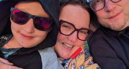 Elena, her husband and young son cuddled together in a selfie, with blue sky behind them.