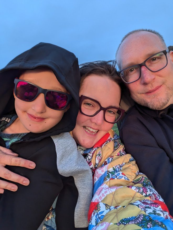 Elena, her husband and young son cuddled together in a selfie, with blue sky behind them.