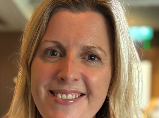 A head and shoulders shot of a white woman in a white top smiling at the camera.