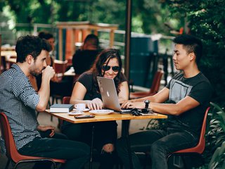 Three friends sat around an outdoor table, surrounded by foliage, drinking coffee and working on a laptop.