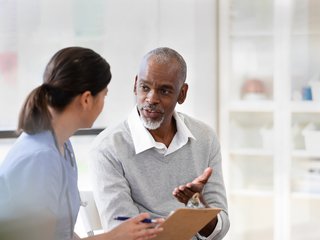 An older man wearing a white shirt and grey jumper is in consultation with his doctor. He is making eye contact and looks like he is asking a question.