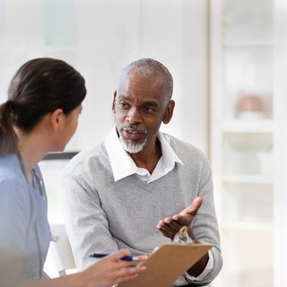 An older man wearing a white shirt and grey jumper is in consultation with his doctor. He is making eye contact and looks like he is asking a question.
