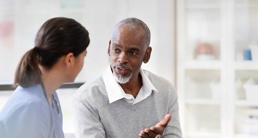 An older man wearing a white shirt and grey jumper is in consultation with his doctor. He is making eye contact and looks like he is asking a question.