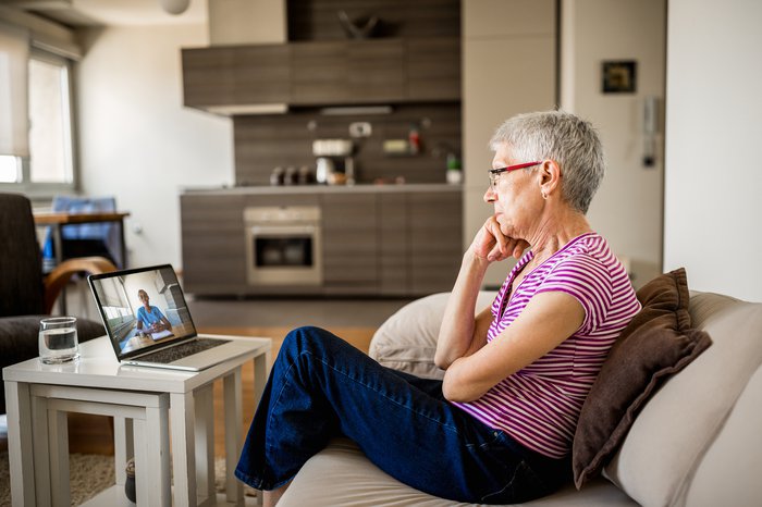 A woman with short grey hair sitting on the sofa in a living room, on a video call with someone on her laptop.