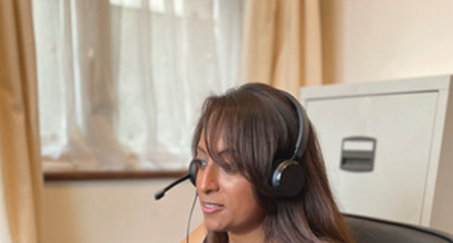 A woman takes a call on a headset as she works from home, looking at the computer in front of her. She's seated at a leather office chair with a filing cabinet and window behind her.