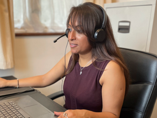 A woman takes a call on a headset as she works from home, looking at the computer in front of her. She's seated at a leather office chair with a filing cabinet and window behind her.