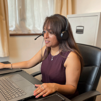 A woman takes a call on a headset as she works from home, looking at the computer in front of her. She's seated at a leather office chair with a filing cabinet and window behind her.