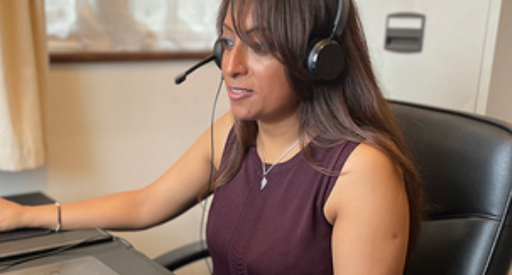 A woman takes a call on a headset as she works from home, looking at the computer in front of her. She's seated at a leather office chair with a filing cabinet and window behind her.