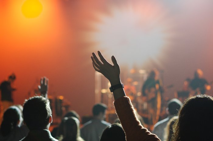 A stock image of a group of people with their hands in the air  enjoying a blurred out band playing in the background.