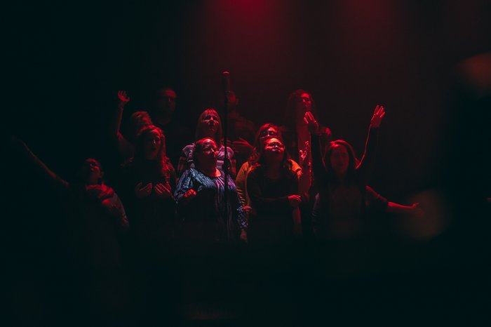 A dark image of a dozen people in a choir singing - their faces lit up in the dark by red spotlights.