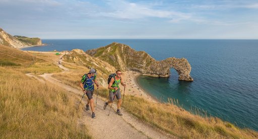 Two men are walking along a coastal path. There is blue sky and Durdle Door beach is in the background.