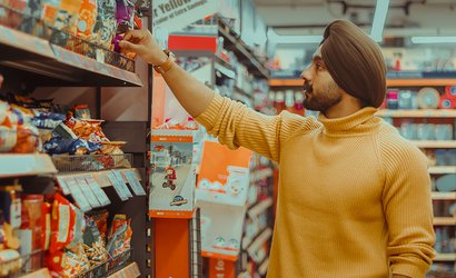 A stock image of a man wearing a turban and a yellow jumper, reaching up to grab a packaged item off a supermarket aisle.