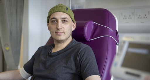 A man having treatment in hospital. He is sitting in a purple hospital bed, wearing a grey t shirt and a woollen beanie hat.