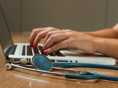 A close up of a person typing on a laptop on a desk. A stethoscope is on the desk to the left of the laptop.