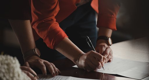 A close up of two people leaning on a desk, one of whom is signing a document