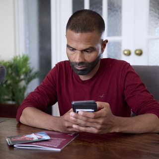 A man sitting at a table, with Blood Cancer UK booklets in front of him. He's holding his phone, as if about to search for something or dial a number.