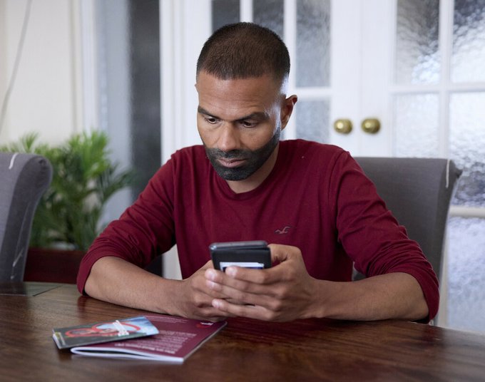 A man sitting at a table, with Blood Cancer UK booklets in front of him. He's holding his phone, as if about to search for something or dial a number.