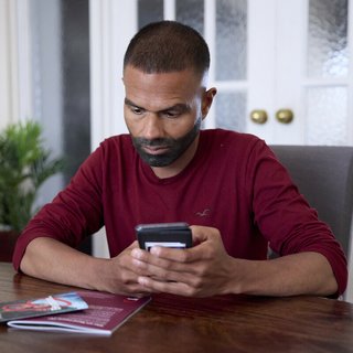 A man sitting at a table, with Blood Cancer UK booklets in front of him. He's holding his phone, as if about to search for something or dial a number.