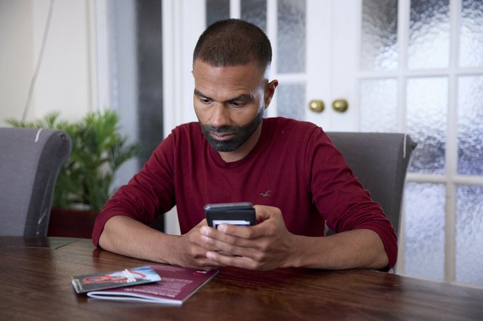 A man sitting at a table, with Blood Cancer UK booklets in front of him. He's holding his phone, as if about to search for something or dial a number.