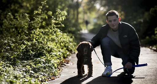 Teenage Blood Cancer UK ambassador kneeling down next to small dog on a concrete path next to bushes.