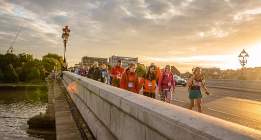 A group of walkers taking part in a charity walk cross the river Thames. The sun is setting and they are smiling.