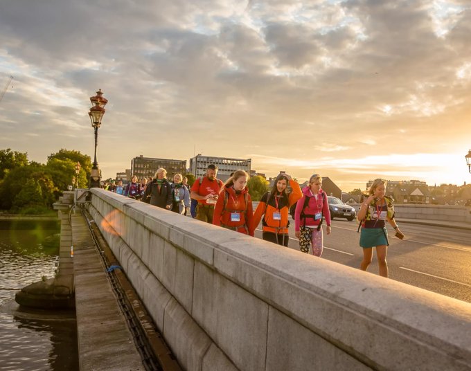 A group of walkers taking part in a charity walk cross the river Thames. The sun is setting and they are smiling.