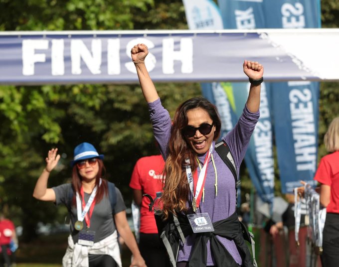 A woman cheers as she crossed the finish line of the Thames Bridges Trek. She has her arms lifted in celebration. She's wearing sunglasses, walking clothes and a charity lanyard.