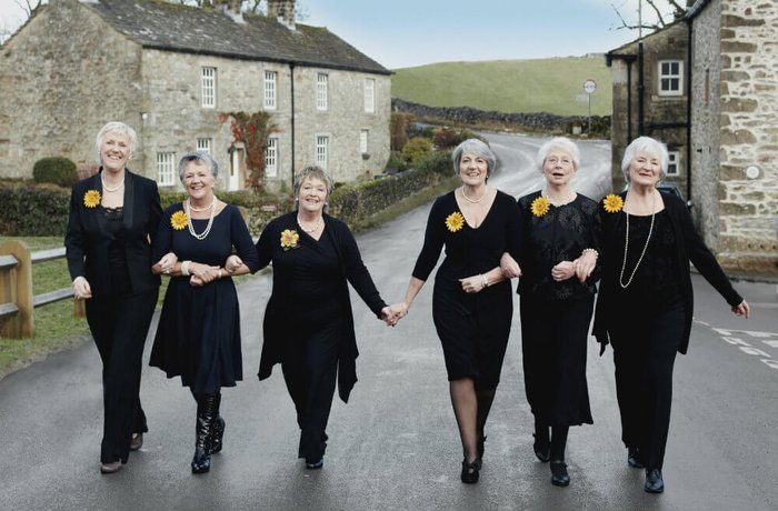 The Calendar Girls smiling and linking arms walking down a road in Yorkshire between two rows of houses.