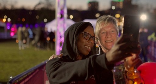 Two women take a selfie at a walk for charity. A black woman wearing glasses and a hoody is taking the photo and a white woman with light grey hair is standing next to her. They are smiling at the camera and there is a park and lights in the background.