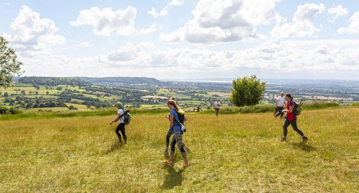 A team of walkers are walking across the peak district. It is a sunny day and there is a panoramic view in the distance.