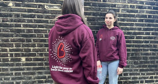 Two people standing outside showing off their Blood Cancer UK, Walk of Light branded hoodies. The hoodies are maroon with hearts and stars.