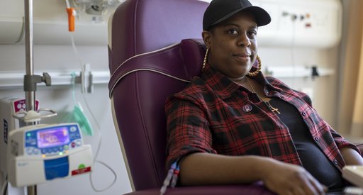 A woman sitting in a hospital receiving intravenous treatment.   She's sitting in a chair, with a thin plastic tube coming out from under her rolled up shirt sleeve. There's an IV stand next to her.