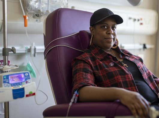 A woman sitting in a hospital receiving intravenous treatment.   She's sitting in a chair, with a thin plastic tube coming out from under her rolled up shirt sleeve. There's an IV stand next to her.