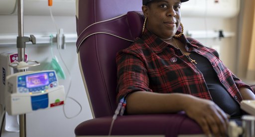 A woman sitting in a hospital receiving intravenous treatment.   She's sitting in a chair, with a thin plastic tube coming out from under her rolled up shirt sleeve. There's an IV stand next to her.