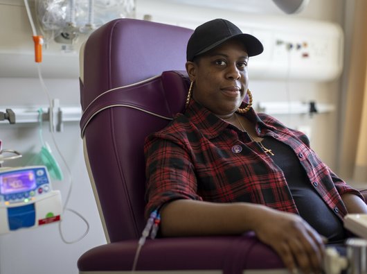 A woman sitting in a hospital receiving intravenous treatment. She's sitting in a chair, with a thin plastic tube coming out from under her rolled up shirt sleeve. There's an IV stand next to her.